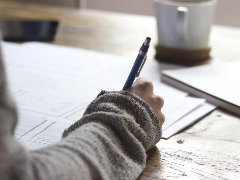 Une main qui écrit au stylo sur des feuilles posées sur une table de bois